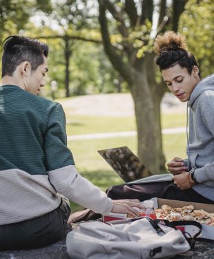Diverse students eating pizza while studying with laptop in park