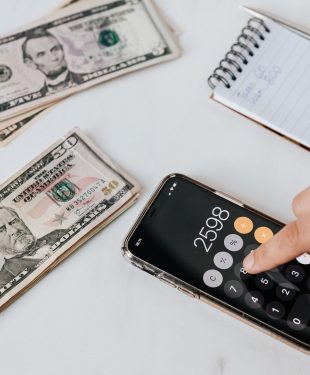 Crop unrecognizable financier using calculator on smartphone near dollar banknotes
